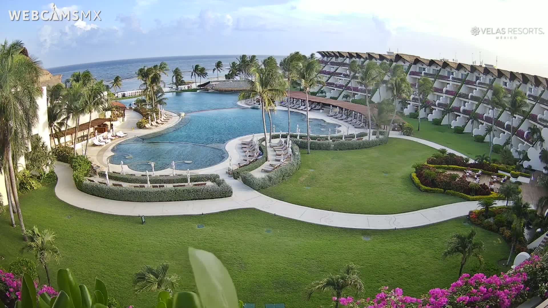 An aerial view of a resort on a sunny day shows a large, multi-level pool complex surrounded by lush green lawns, palm trees, and a multi-story building with balconies overlooking the ocean.