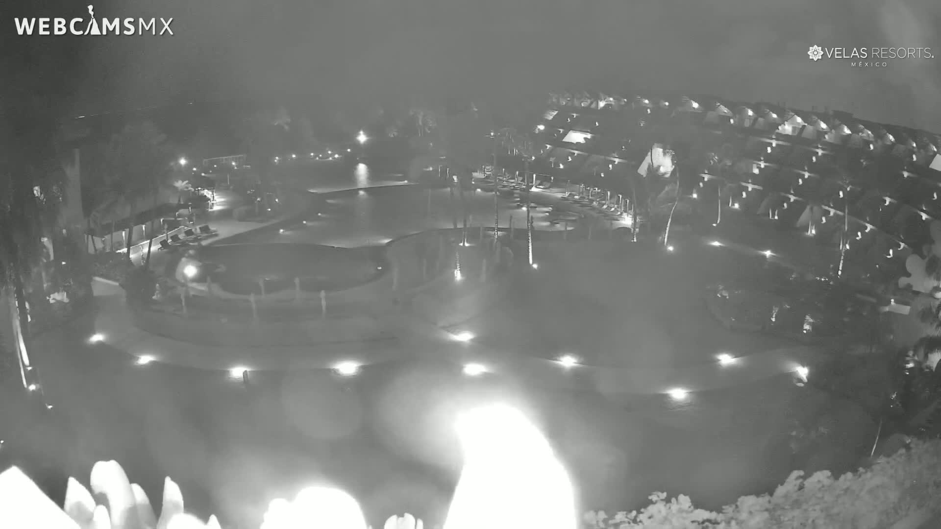 A nighttime aerial view of a resort with swimming pools and low-light illumination, under hazy or foggy conditions.