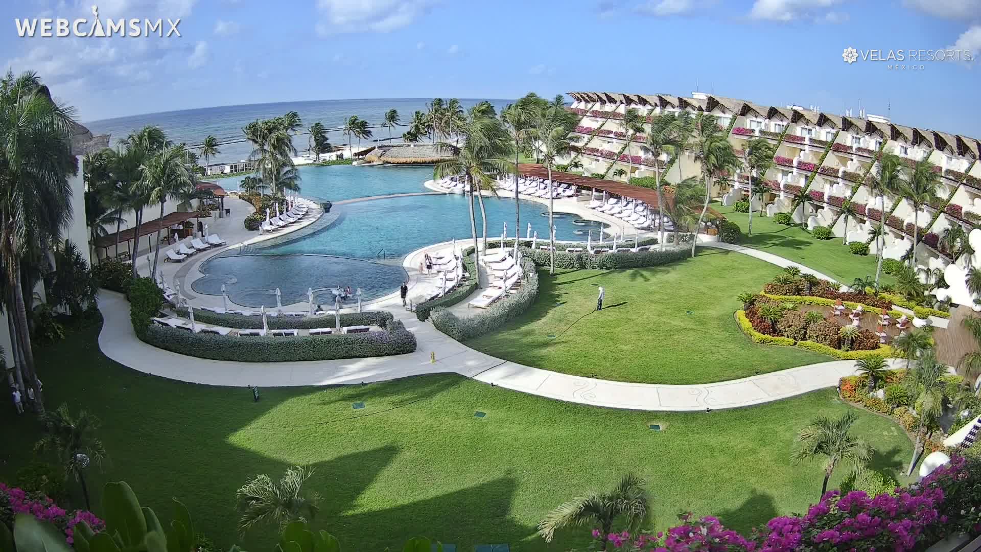A resort with a large pool and ocean view, under sunny skies with a slight ocean breeze.