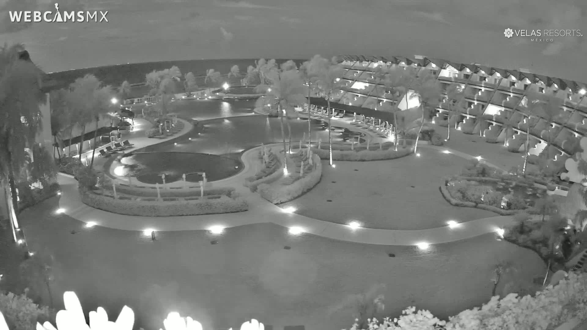 A nighttime aerial view shows a resort with a winding pool, surrounding landscaping, and a multi-story building under a partly cloudy sky.