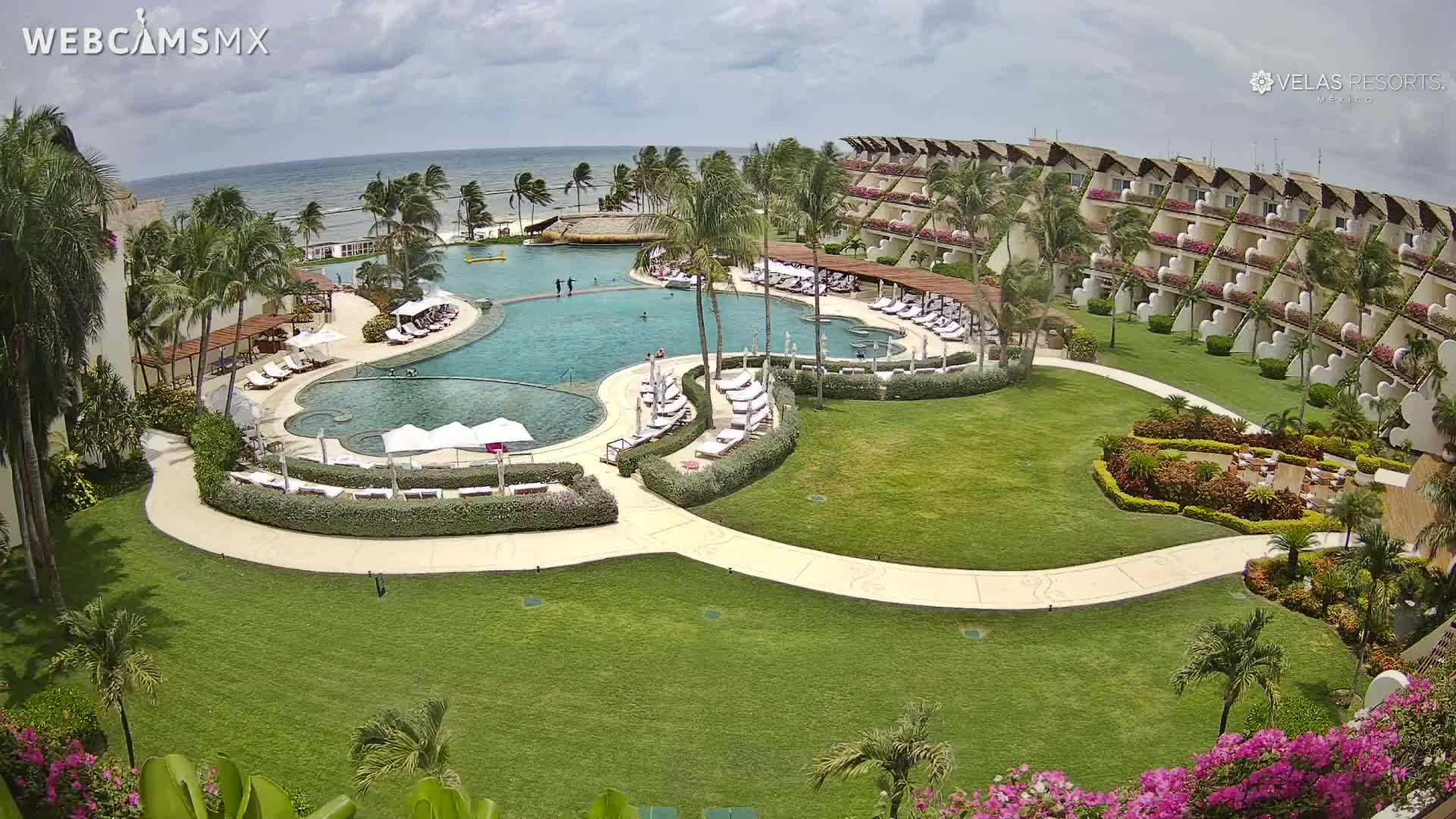 A resort's pool area with a large, freeform pool surrounded by lounge chairs and lush landscaping, situated next to a multi-story building under a partly cloudy sky.