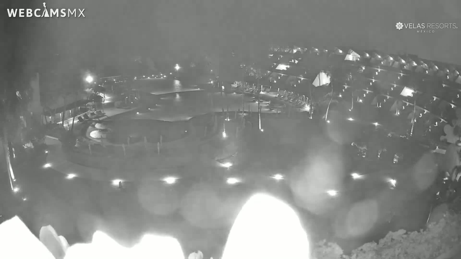 A nighttime, blurry aerial view of a resort with a large pool and surrounding buildings, under slightly overcast conditions.