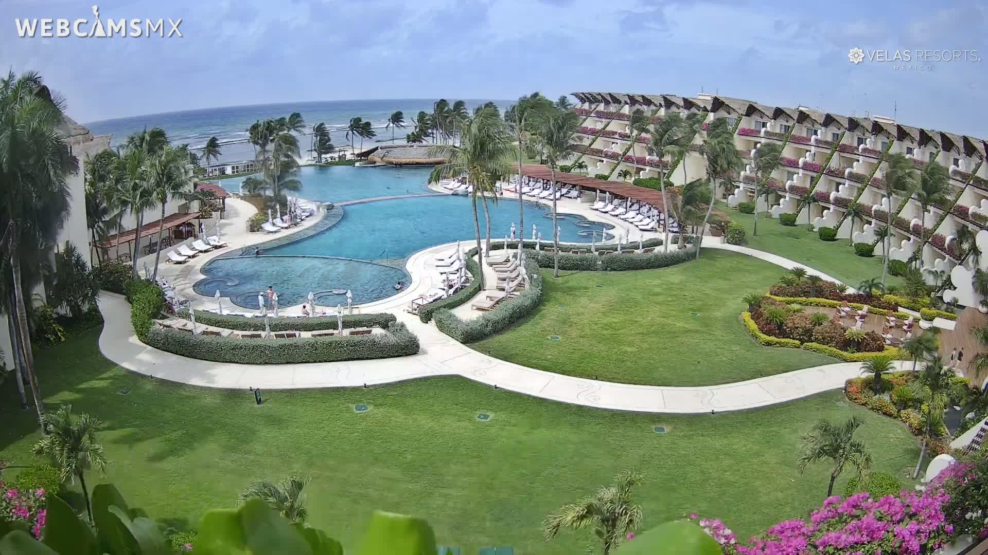 An aerial view of a resort on a partly cloudy day shows a large, curved swimming pool surrounded by lounge chairs and landscaping, with a beachfront building complex and ocean visible in the background.