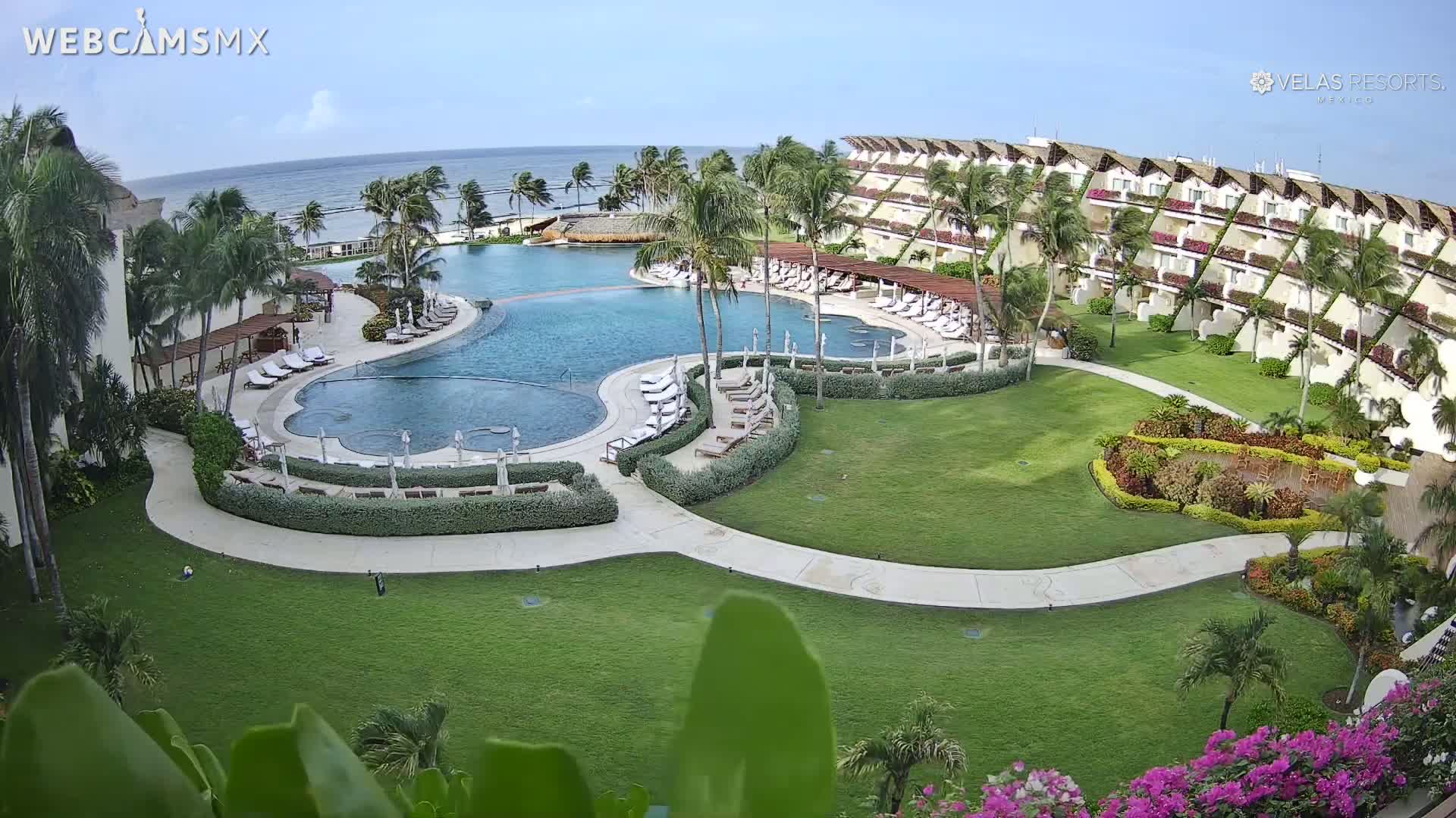 An aerial view of a resort with a large, curved swimming pool surrounded by lush landscaping, palm trees, and a multi-story building overlooking a calm ocean under a clear sky.