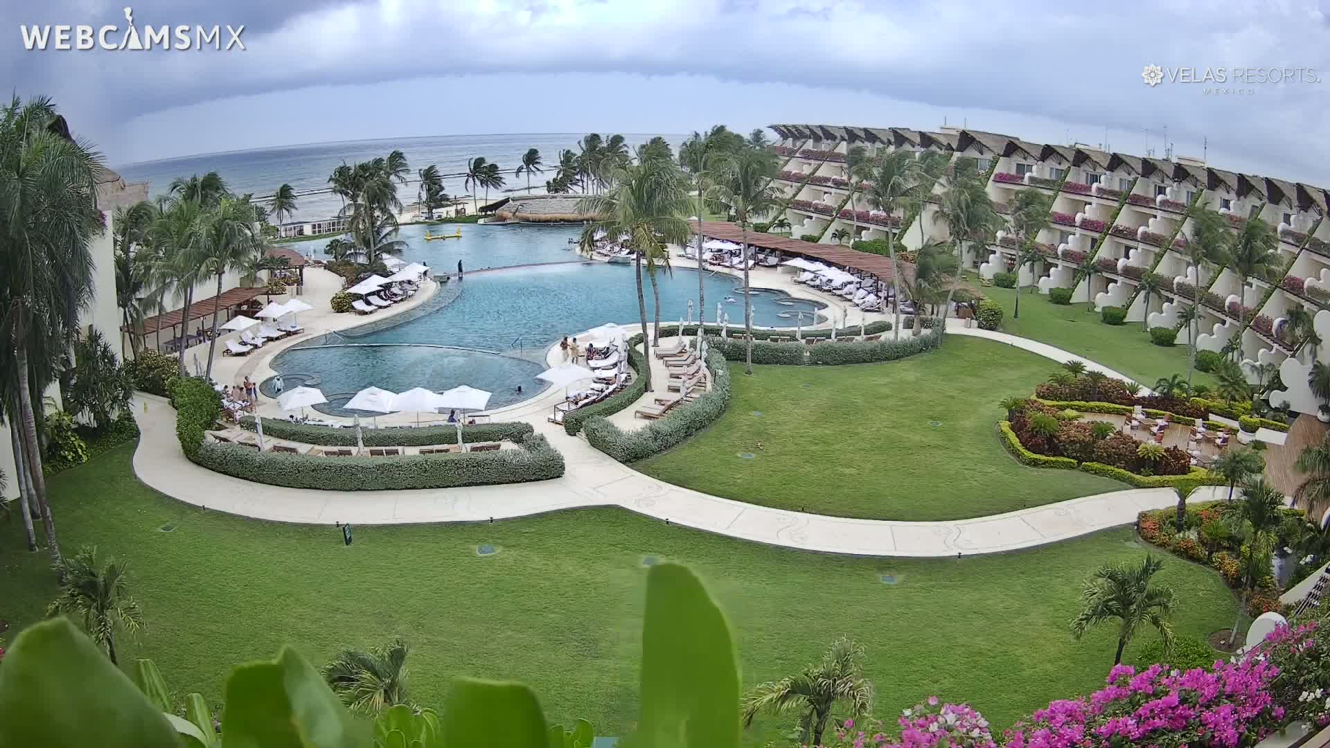 An aerial view shows a resort pool and surrounding buildings on a partly cloudy day, with a calm ocean visible in the background.