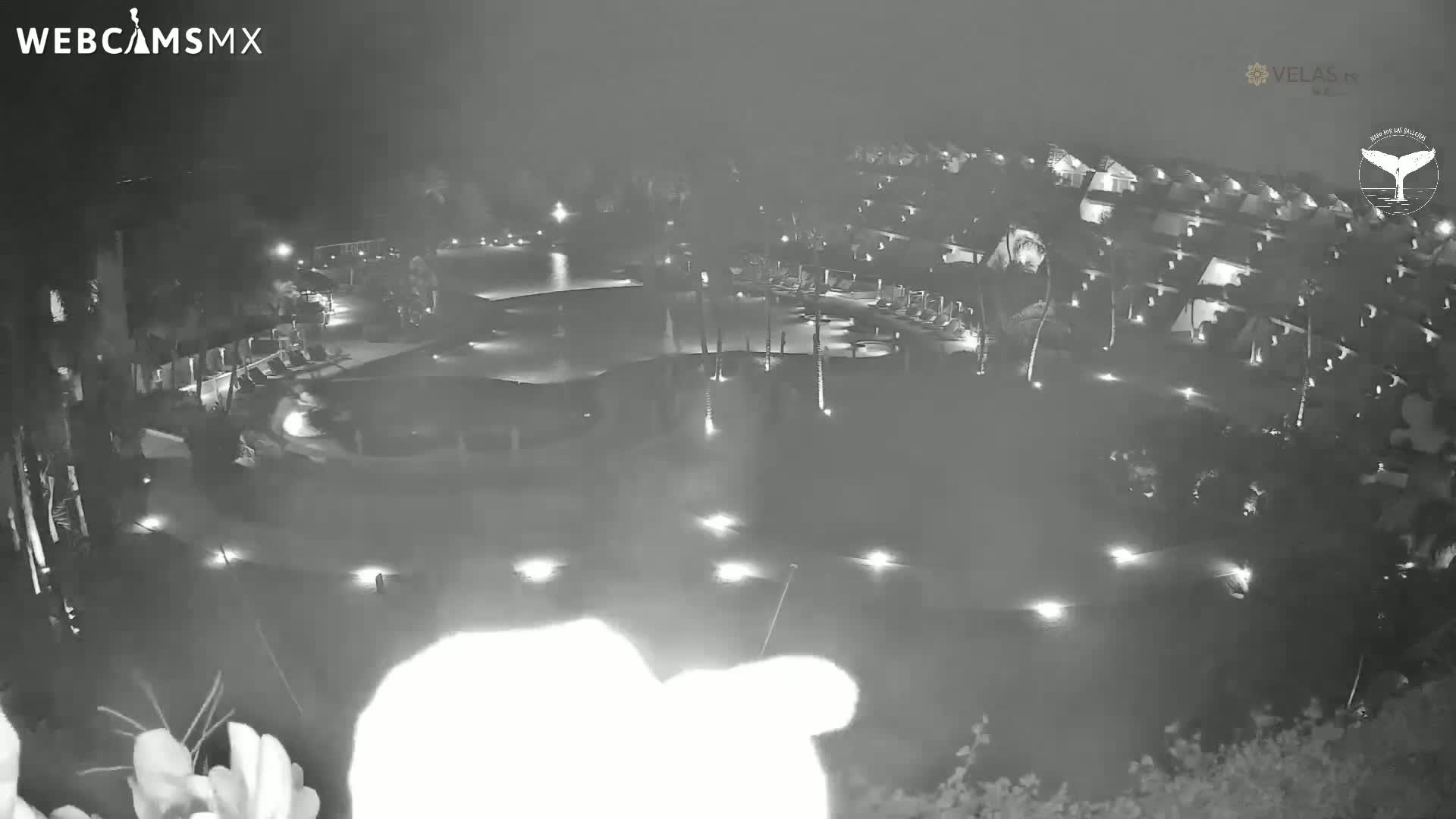 A nighttime, low-light aerial view shows a resort with a large pool and numerous buildings illuminated by scattered lights.