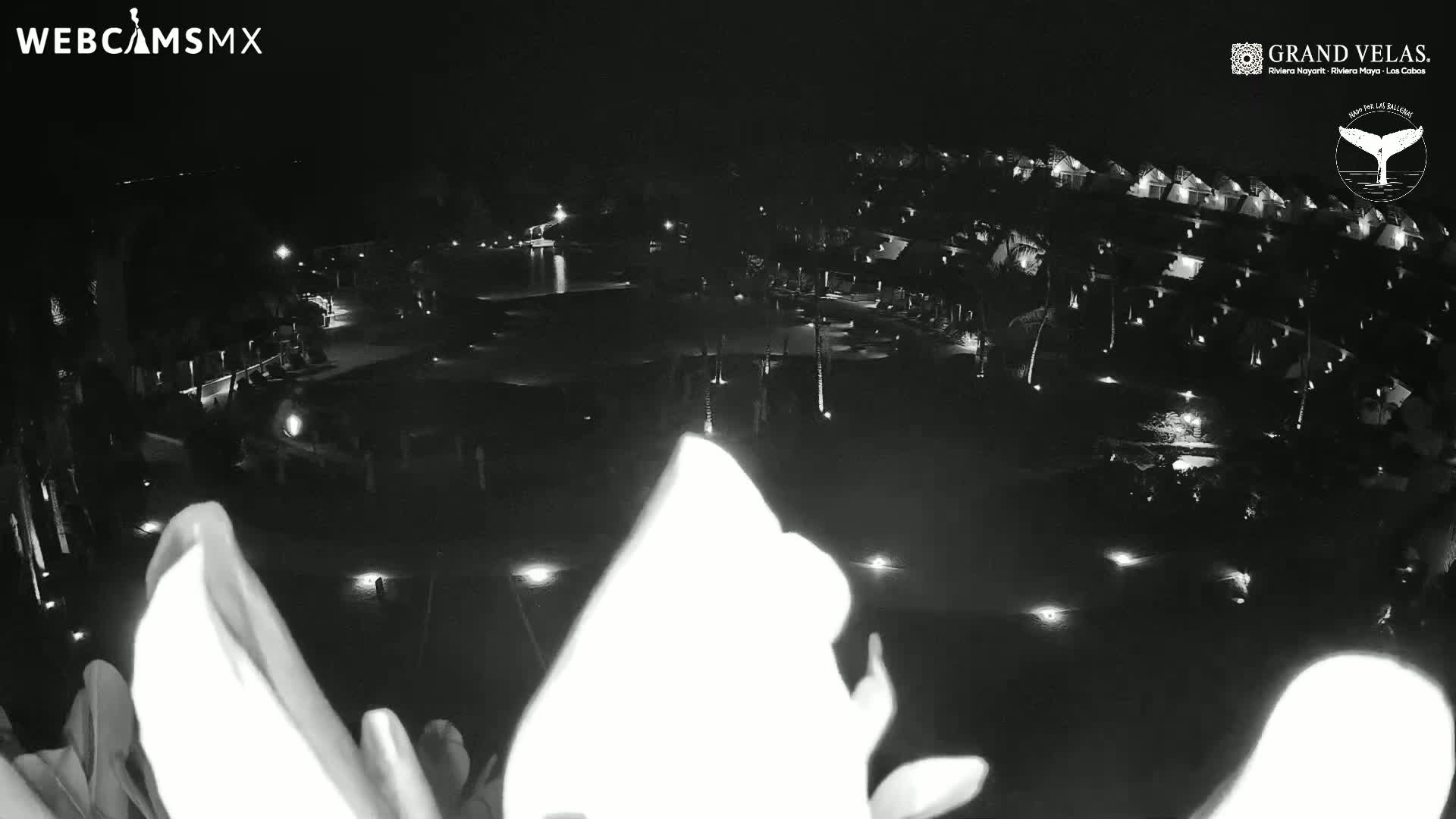 A nighttime, low-light aerial view shows a resort with a large pool and numerous buildings illuminated by scattered lights.