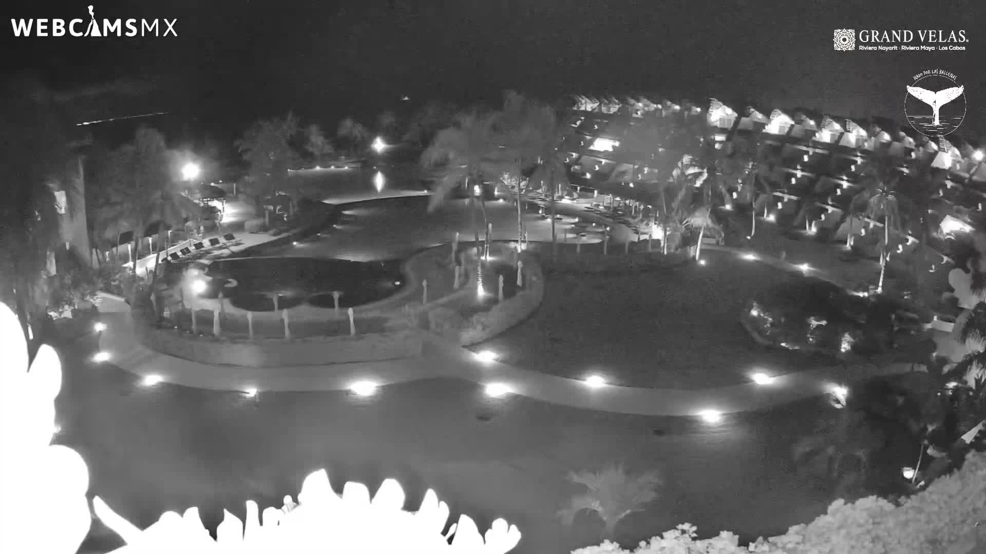 A nighttime view reveals an illuminated resort complex with multiple swimming pools, palm trees, and buildings, all under clear skies.