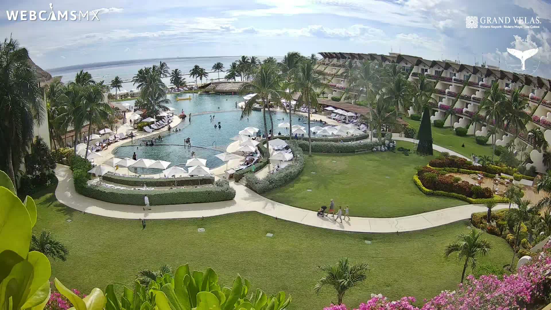 A vibrant tropical resort scene is depicted on a partly cloudy day, featuring a large, winding swimming pool with numerous guests, white umbrellas, and lounge chairs, surrounded by lush green lawns, swaying palm trees, a multi-story hotel building with many balconies, and the calm ocean stretching into the distance.