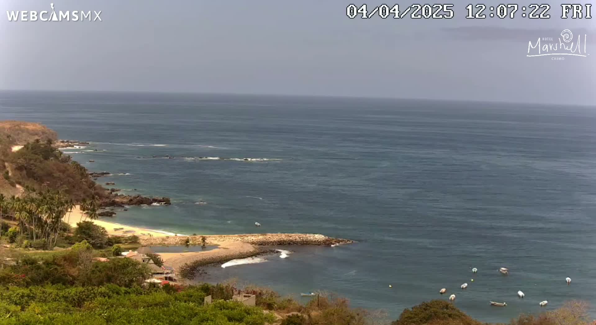 A high-angle view shows a calm ocean with several small boats, a sandy beach, rocky coastline, and lush green vegetation under a hazy sky.