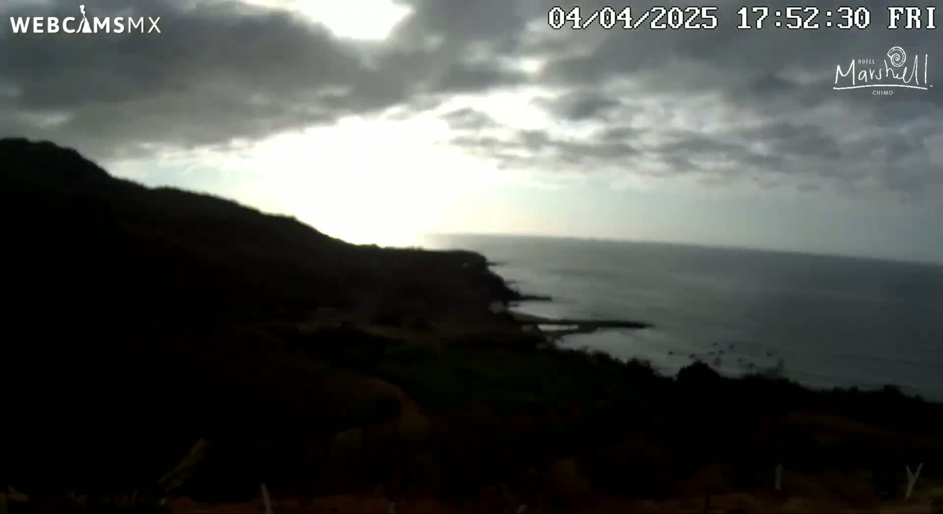 A partly cloudy sunset over a dark, mountainous coastline and a calm ocean.