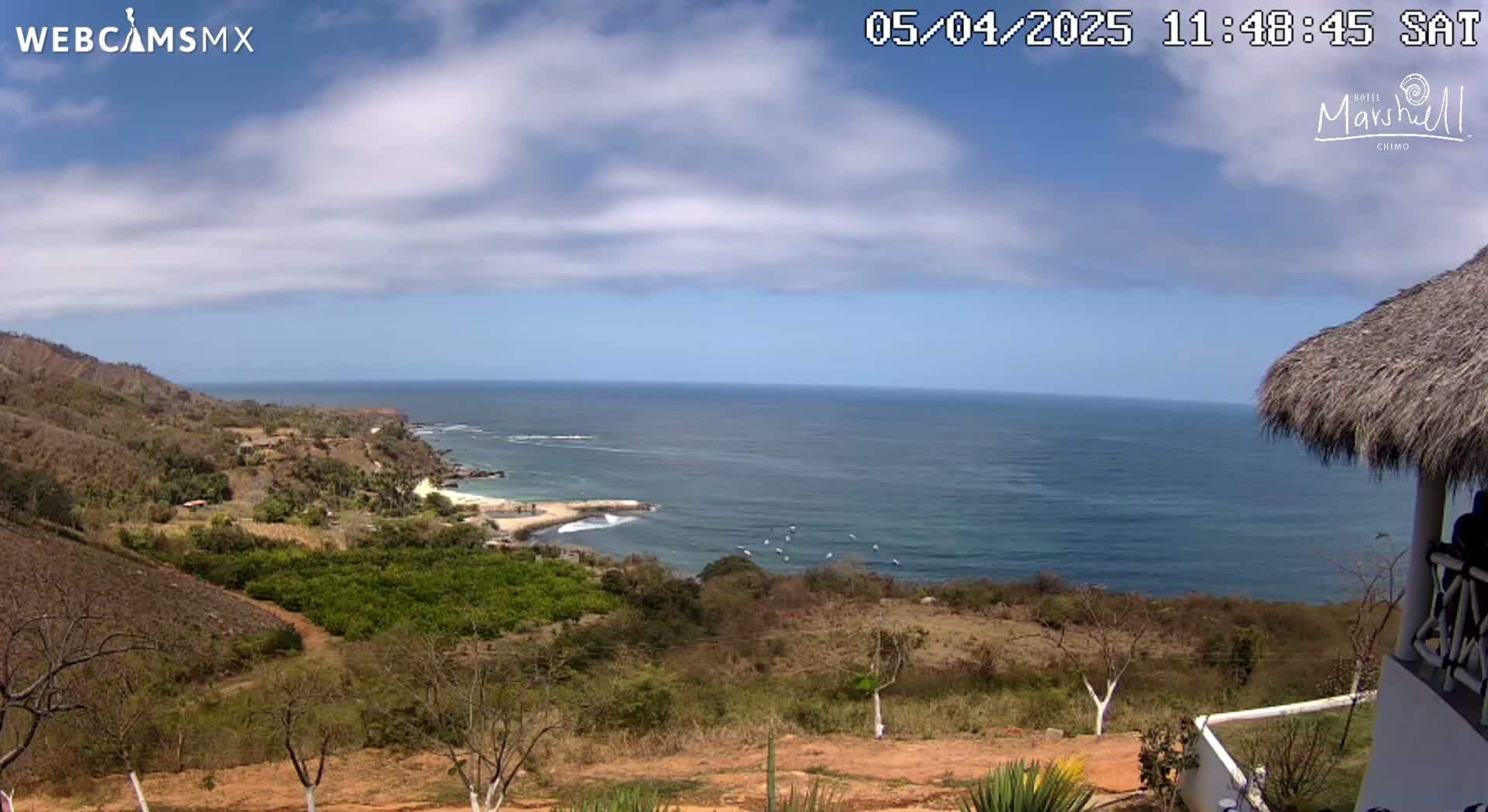 A partly cloudy day overlooking a tranquil ocean scene from a hillside, featuring a small beach and a thatch-roofed structure.