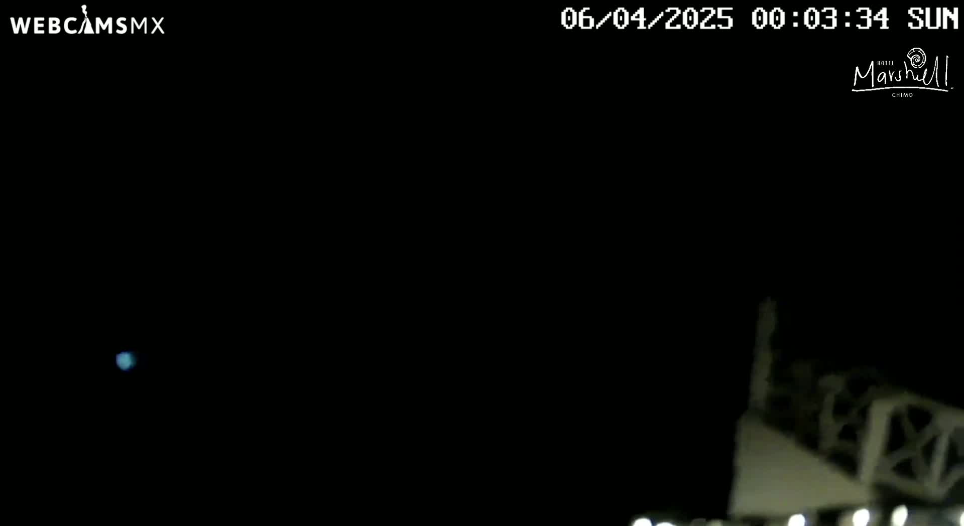 A dark nighttime scene shows a dimly lit structure in the lower right corner, with a faint light visible in the distance under a clear, dark sky.