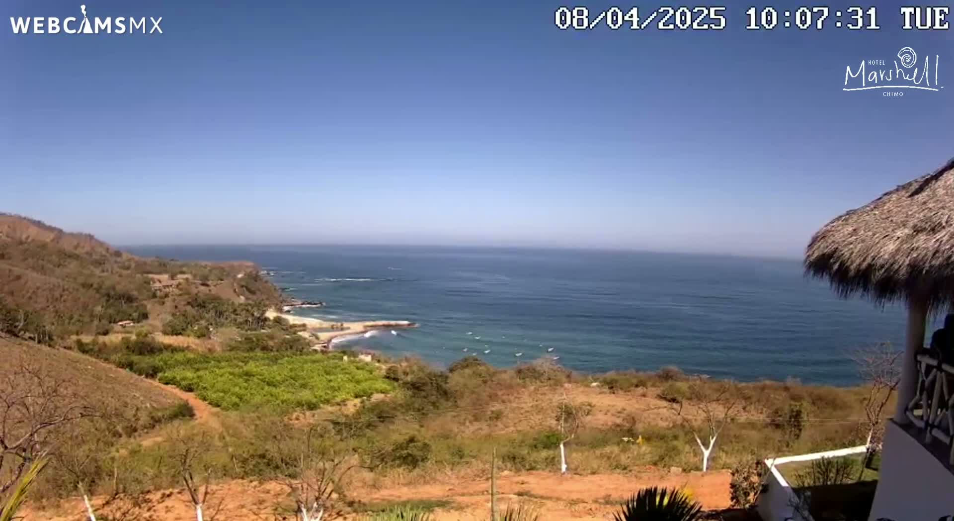 A high-angle view shows a tranquil ocean scene under a clear blue sky, with dry, brown hillsides and sparse vegetation leading down to a sandy cove.