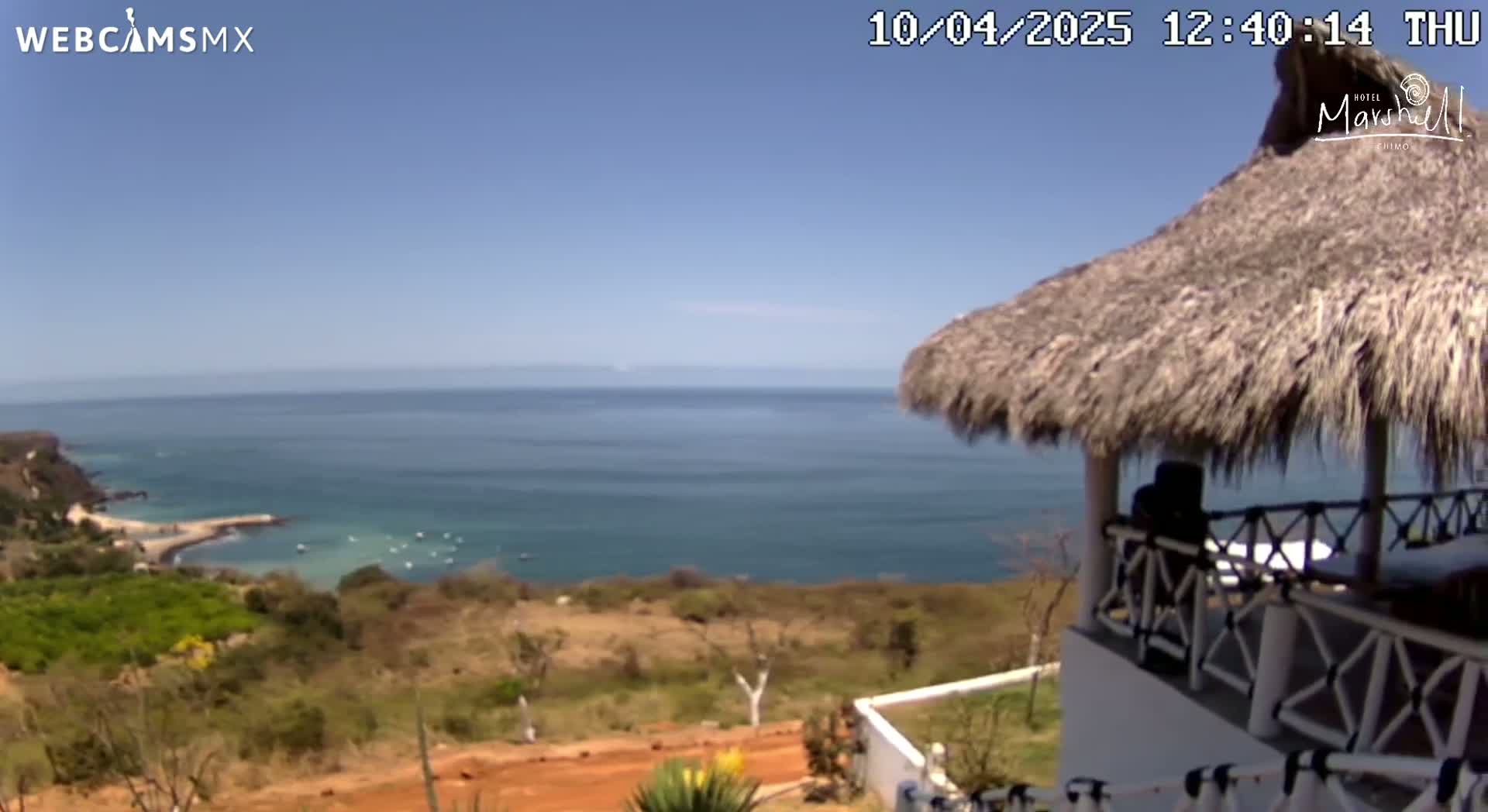 A thatched-roof structure overlooks a calm ocean on a sunny day, with a coastal landscape visible in the foreground.