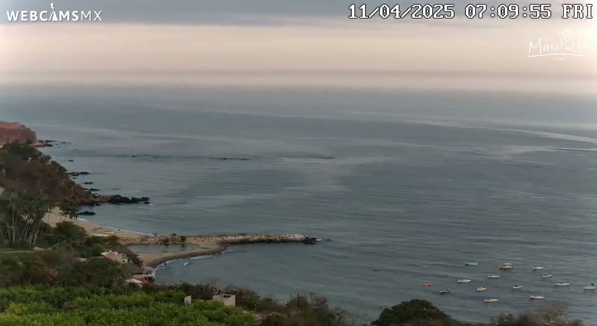 An elevated view shows a calm, hazy ocean with several small boats near a rocky coastline, featuring a small beach and vegetation-covered hills.