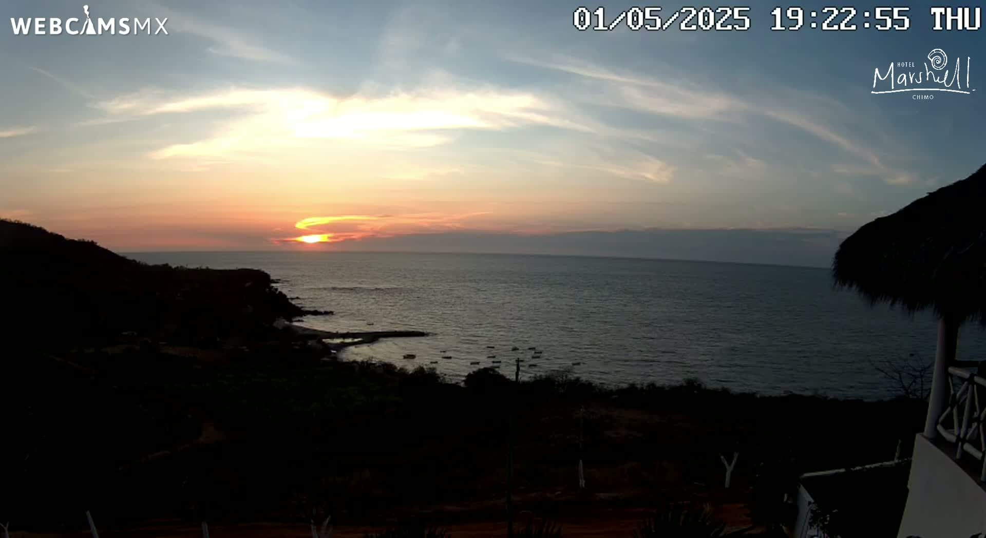 A sunset over a calm ocean is viewed from a hilltop, with a portion of a thatched roof structure visible in the foreground.