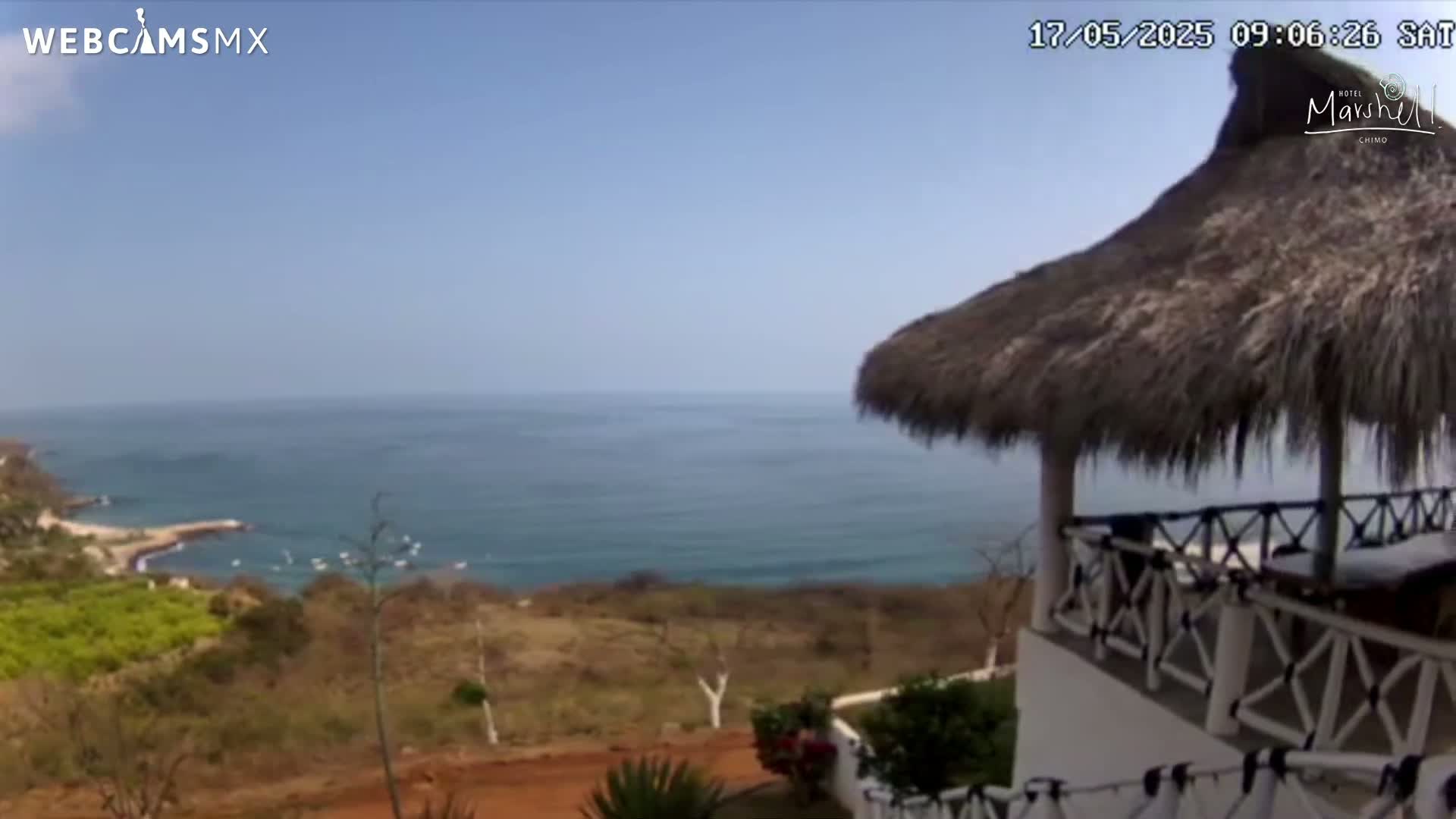 A partly sunny day overlooking a calm ocean from a seaside patio with a thatched roof.