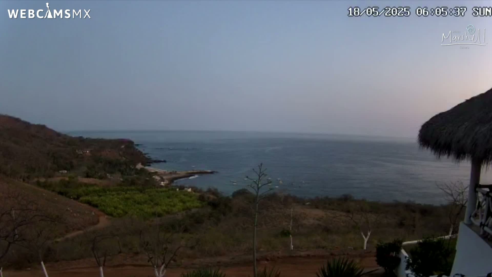 A tranquil coastal scene at dawn or dusk shows a calm ocean extending to the horizon, with brown hills and sparse vegetation in the foreground, partially obscured by a thatched roof structure.