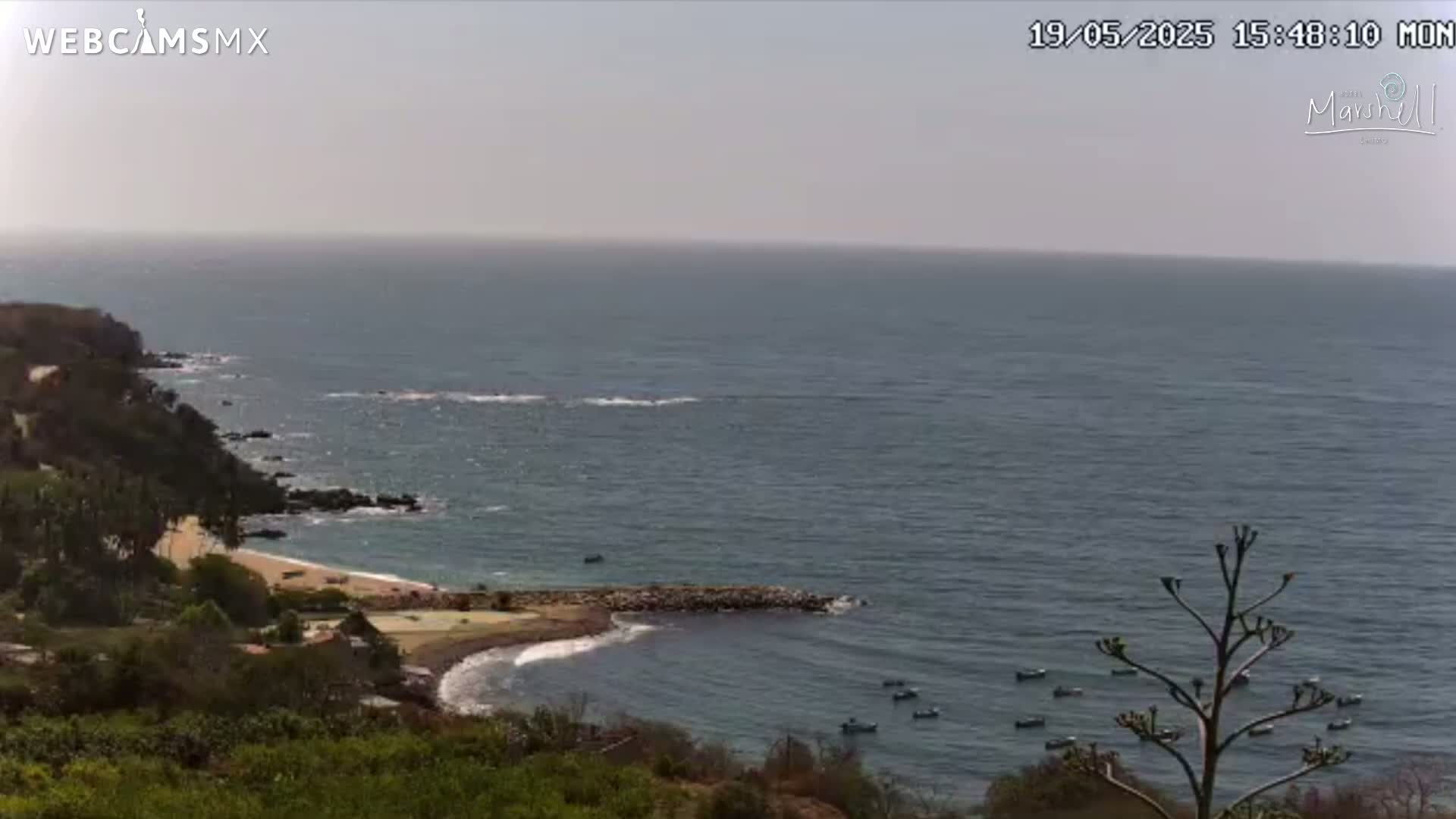 A hazy, sunny day overlooks a calm ocean bay with a small sandy beach, rocky coastline, and several small boats.