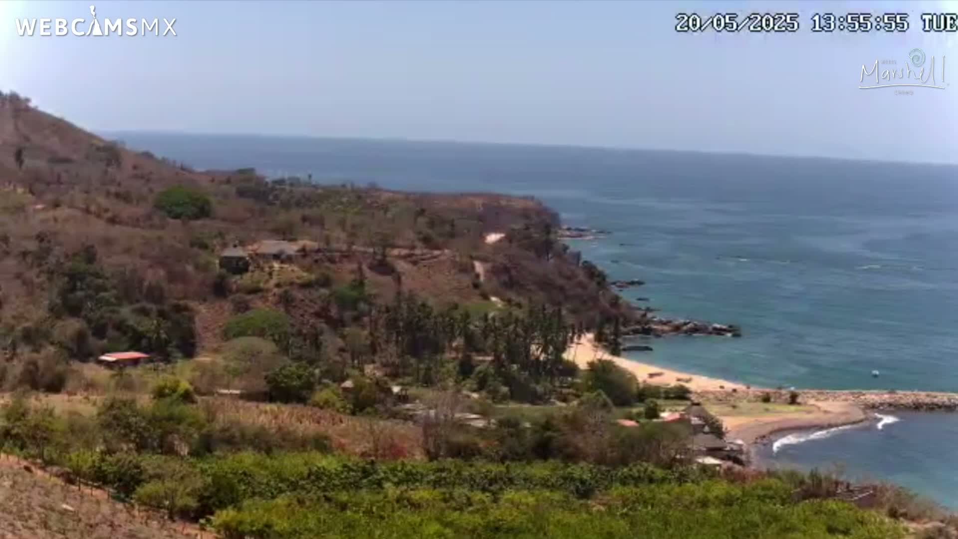 A sunny, high-angle view shows a coastal scene with a sandy beach curving into a rocky cove, backed by sparsely vegetated hills and a calm ocean.