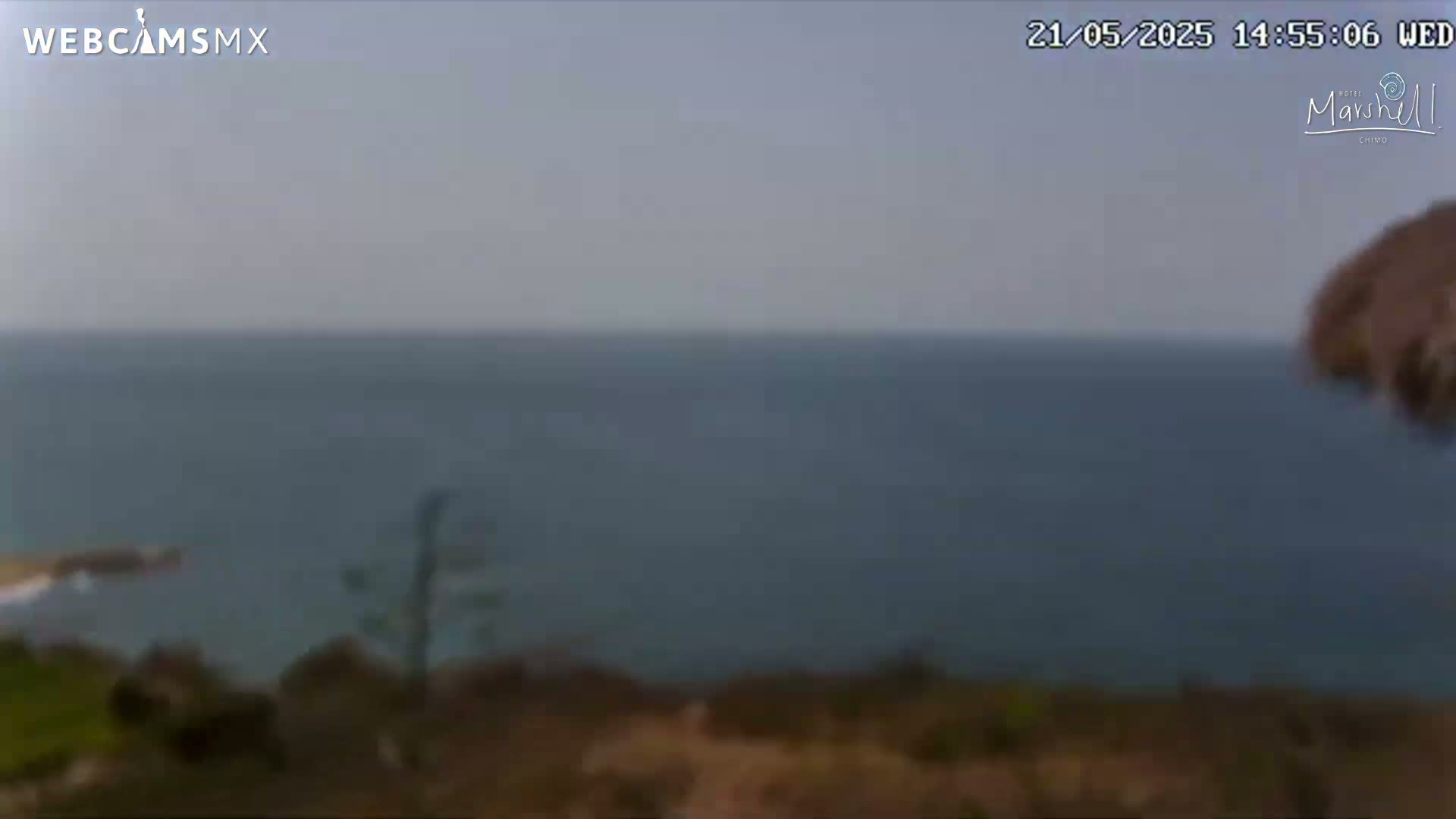 A hazy view of a calm ocean from a coastal vantage point shows brown scrubland in the foreground.