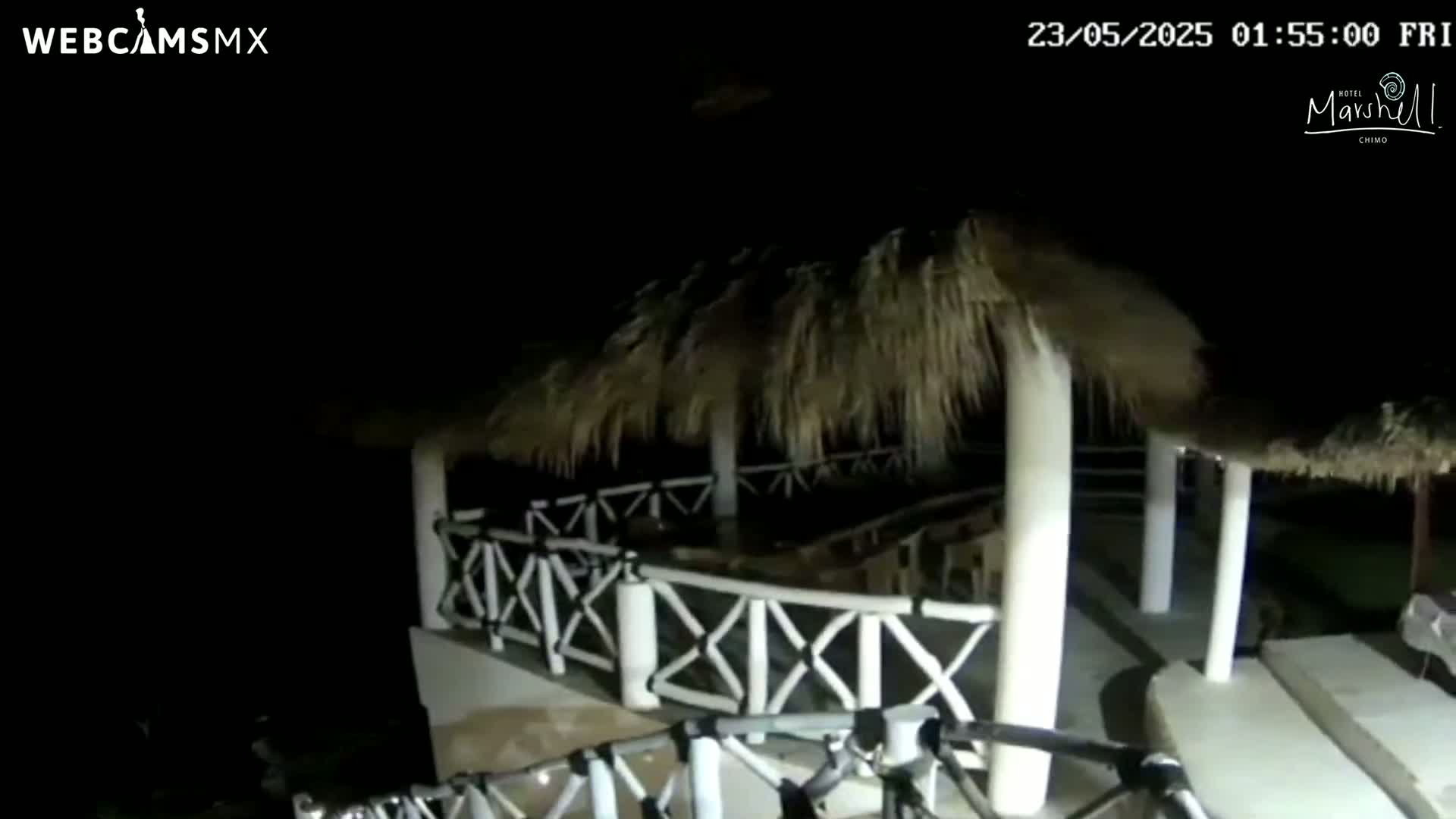 At night, an outdoor patio area with a thatched roof and white railings is dimly lit.