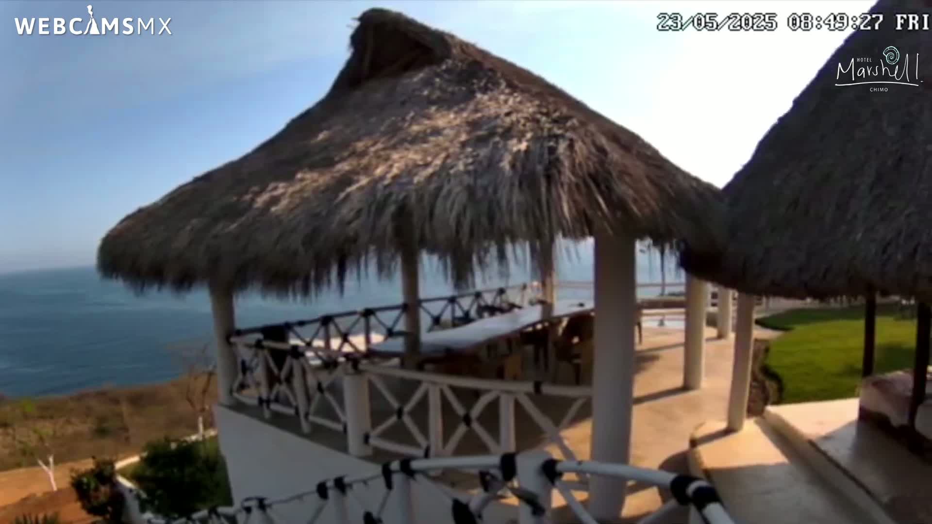 A white structure with a thatched roof overhang and white railing overlooks a calm ocean under a clear, sunny sky.