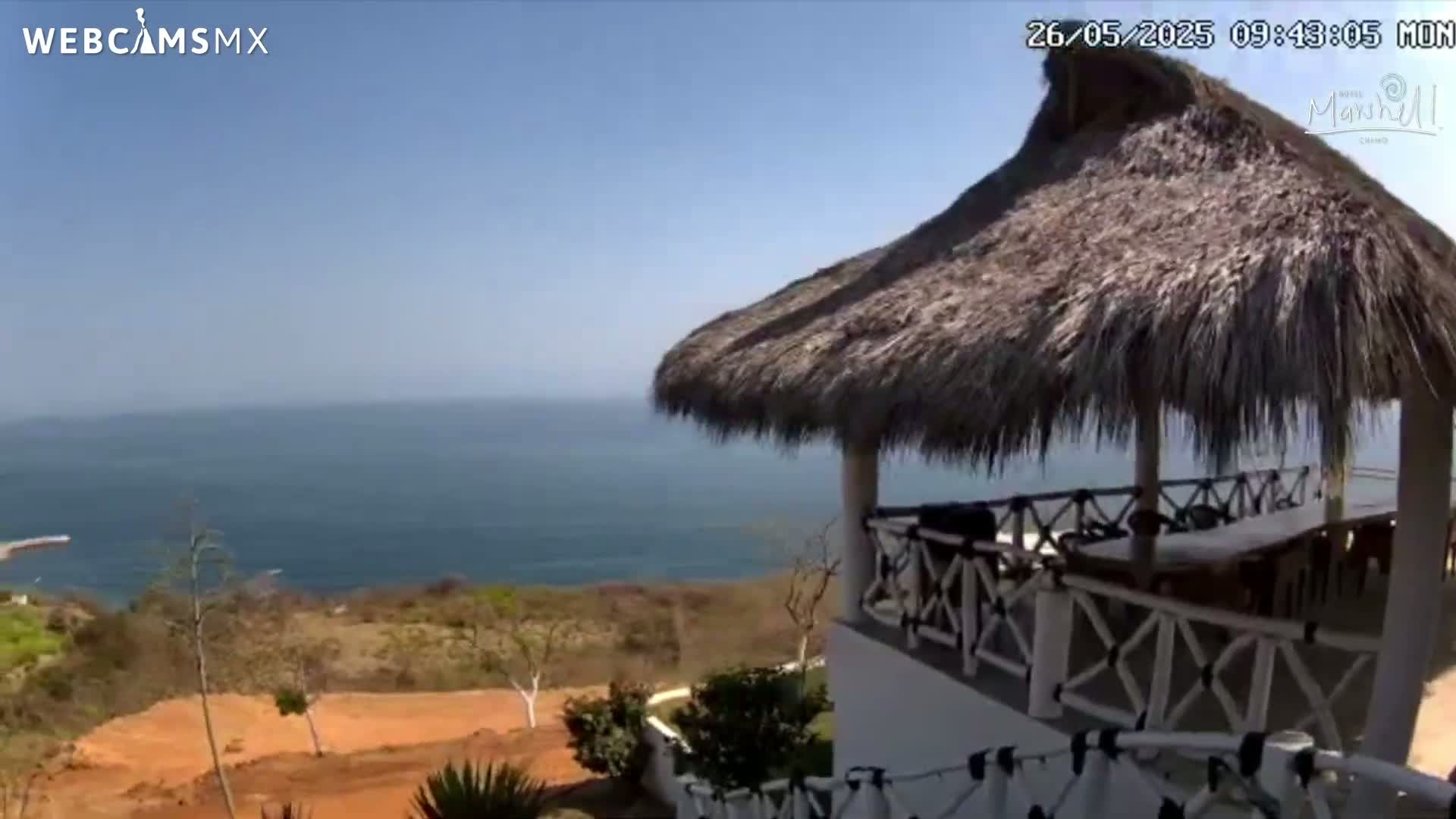 A thatched-roof gazebo overlooks a calm ocean on a sunny day, with brown, scrubby land sloping down to the water.
