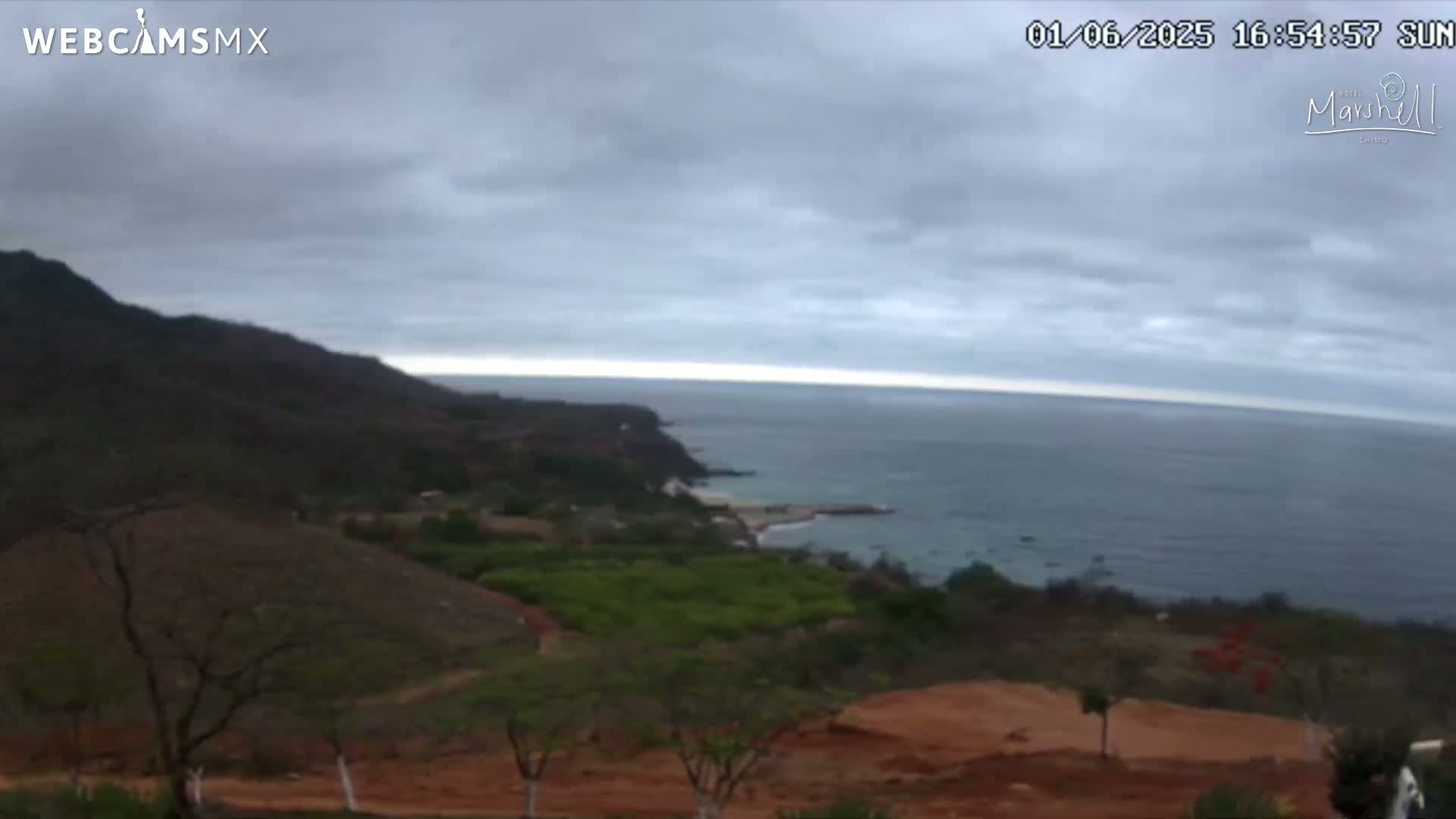 A partly cloudy day overlooks a coastline with dark-green hills, reddish-brown earth, and a calm ocean.