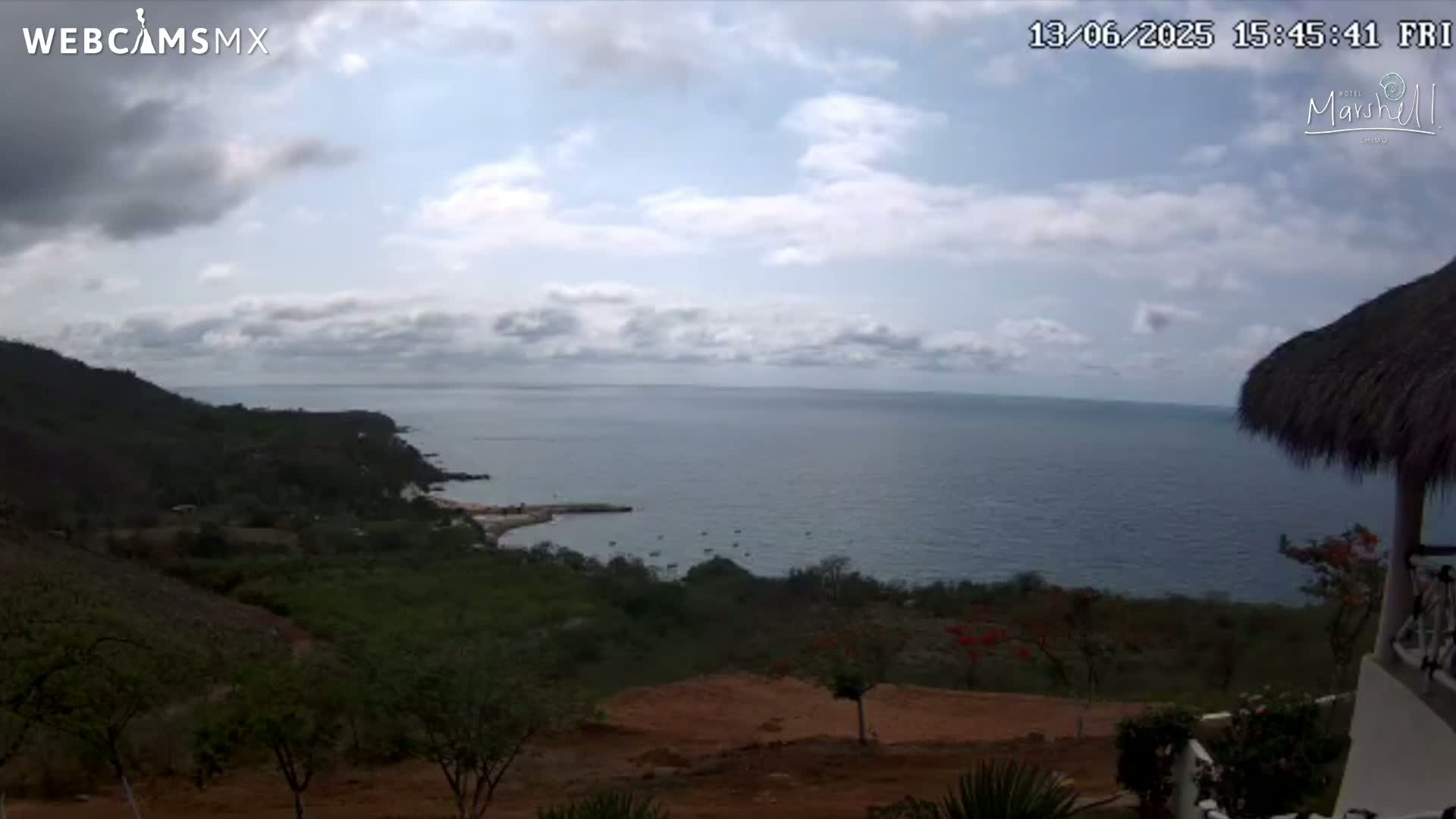 A partly cloudy day overlooks a calm ocean from a hillside covered in green vegetation, with a portion of a thatched-roof structure visible in the bottom right corner.