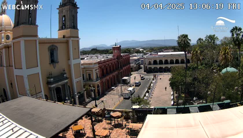 A sunny day overlooking a town square with a large church, colonial-style buildings, and palm trees, viewed from a rooftop patio.