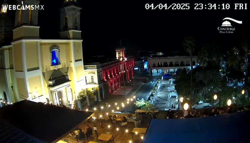 A nighttime view of a city square with a brightly lit cathedral and other buildings, illuminated by string lights and streetlights, under a clear sky.