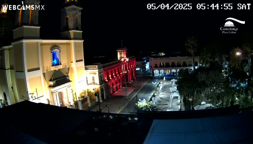 A nighttime view of a town square, illuminated by lights, showing a large cathedral and other buildings, under a clear, dark sky.