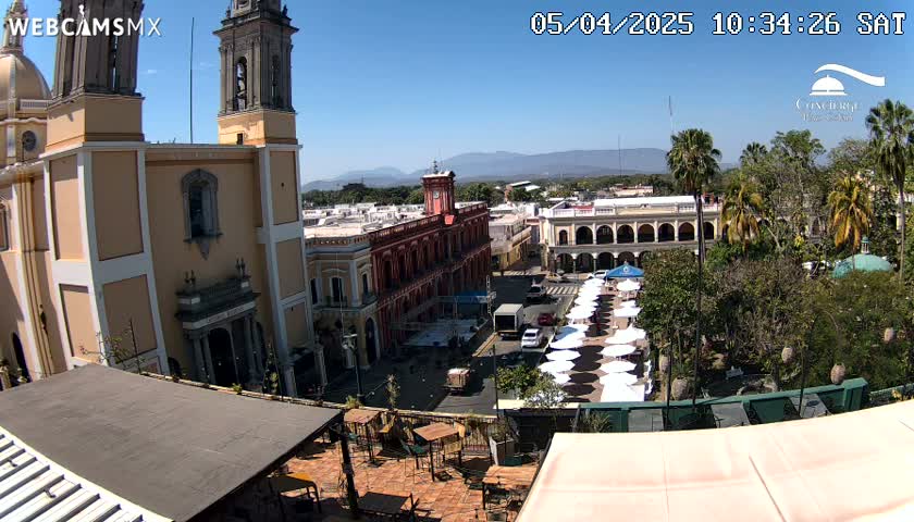 A sunny view of a town square, featuring a large cathedral, colonial-style buildings, numerous white parasols, and palm trees under a clear blue sky.