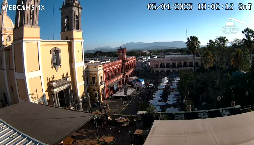 A high-angle, daytime view shows a town square with a large gathering of people under white umbrellas, adjacent to colonial-style buildings, including a prominent yellow church, all under a clear blue sky.