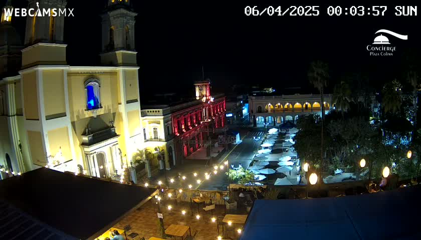 A nighttime view of a town square with a brightly lit cathedral and colonial-style buildings, surrounded by trees and outdoor seating with string lights under a clear night sky.