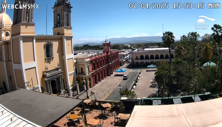 A sunny view of a town square with a large church and colonial-style buildings, surrounded by lush green trees and mountains in the distance.