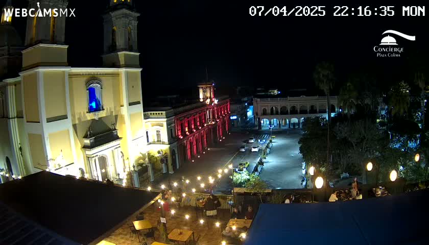 A nighttime view of a town square with a large church and other buildings illuminated, people dining outdoors under string lights.