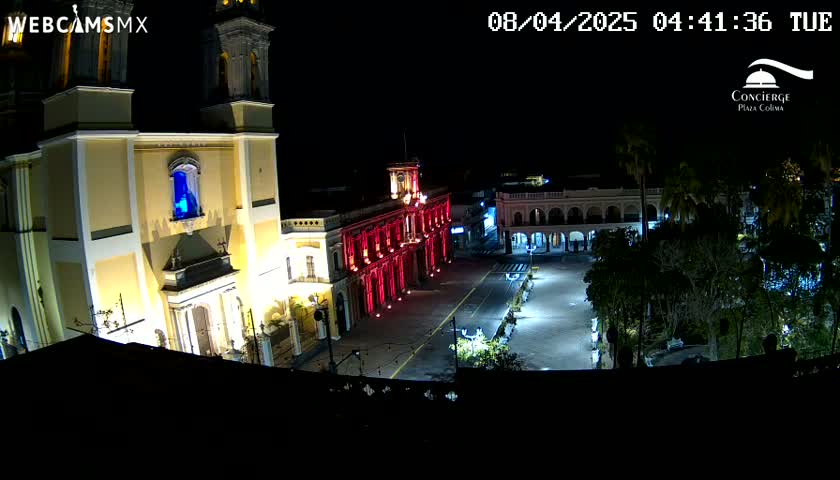 A brightly lit town square at night, featuring a large cathedral and government building illuminated in red, surrounded by trees under a clear, dark sky.