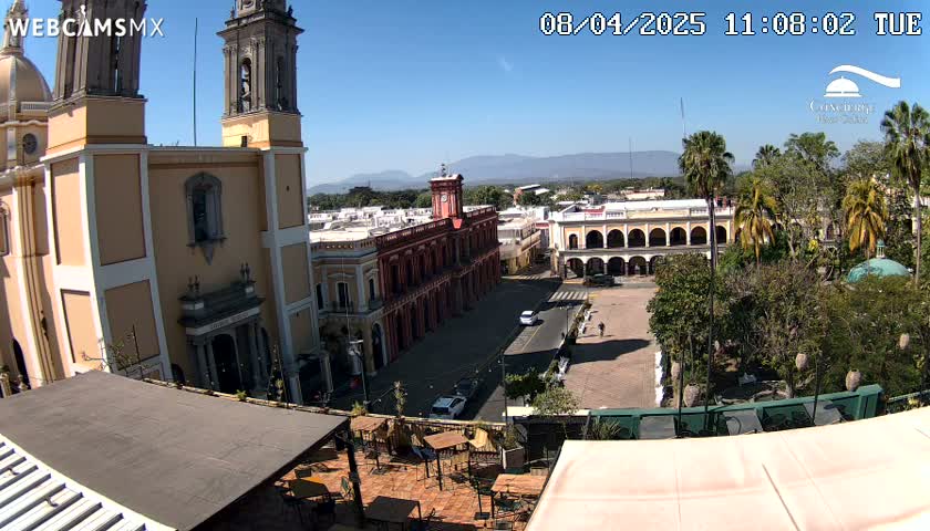 A sunny day reveals a town square with a large cathedral, colonial-style buildings, palm trees, and a rooftop patio with outdoor seating.