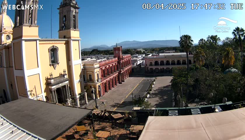A sunny view overlooking a town square, with a large yellow cathedral on the left, red colonial-style buildings across the square, and lush green trees surrounding the plaza.