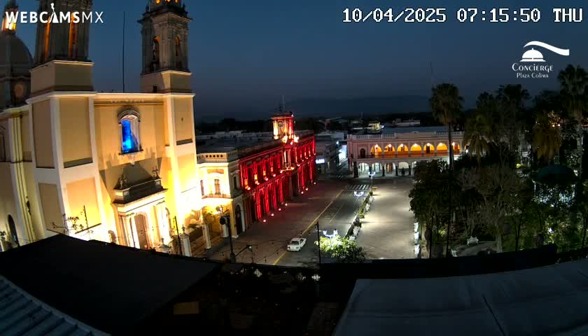 A nighttime view of a town square, illuminated by artificial light, featuring a large yellow church and a red building, surrounded by trees and other buildings.