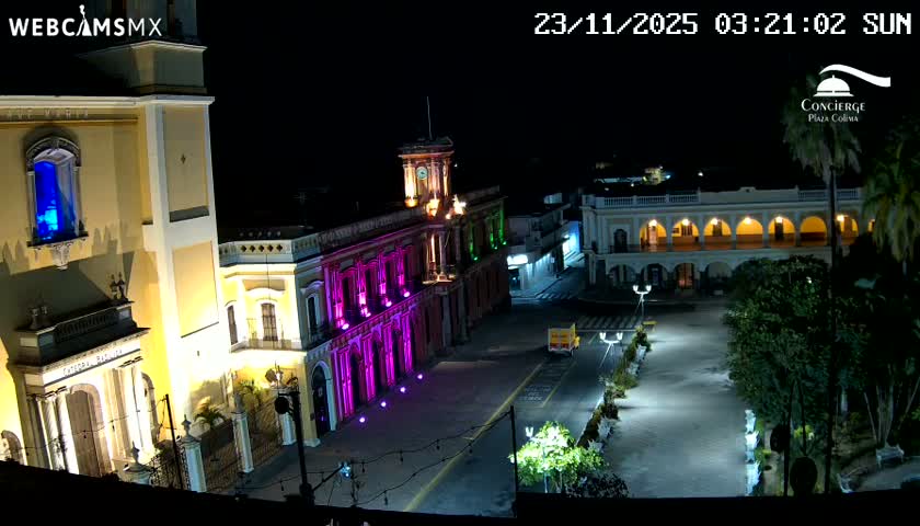 A vibrant city square at night features several historic-looking buildings brightly illuminated in yellow, purple, and green lights, including a grand structure with a blue-lit window, alongside a street with a small truck, under clear conditions.