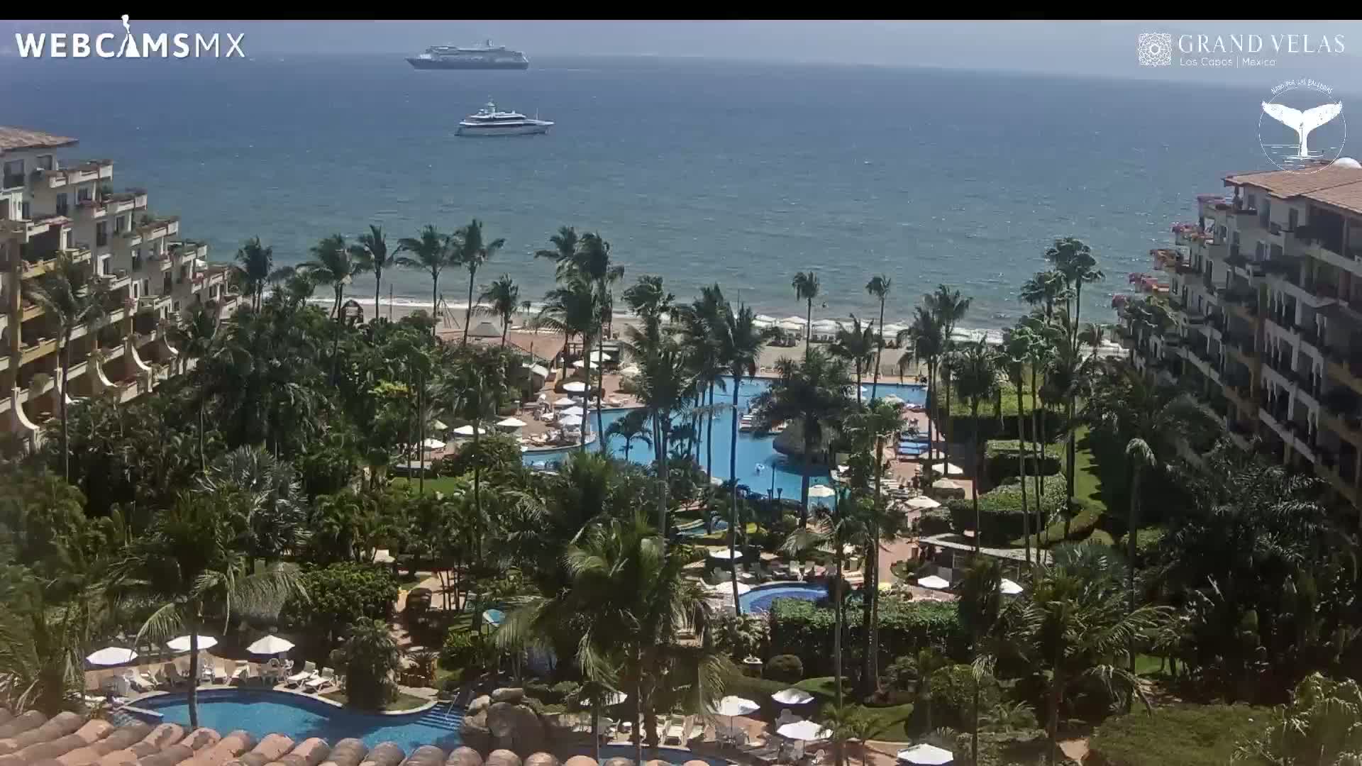 A resort with multiple pools and lush landscaping sits on a coastline, with a calm ocean and two boats visible in the distance under a sunny sky.