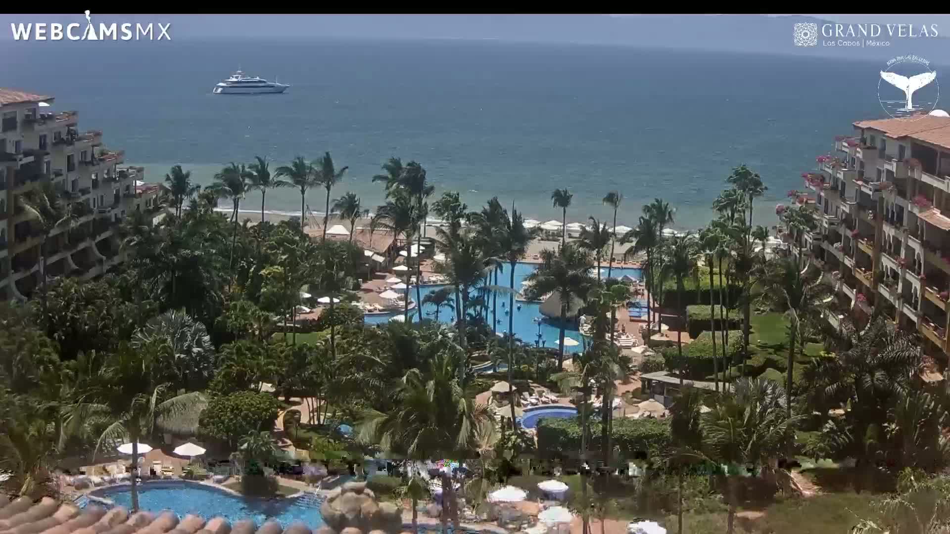 A resort with multiple pools is nestled among palm trees overlooking a calm ocean where a yacht is visible in the distance under a sunny sky.