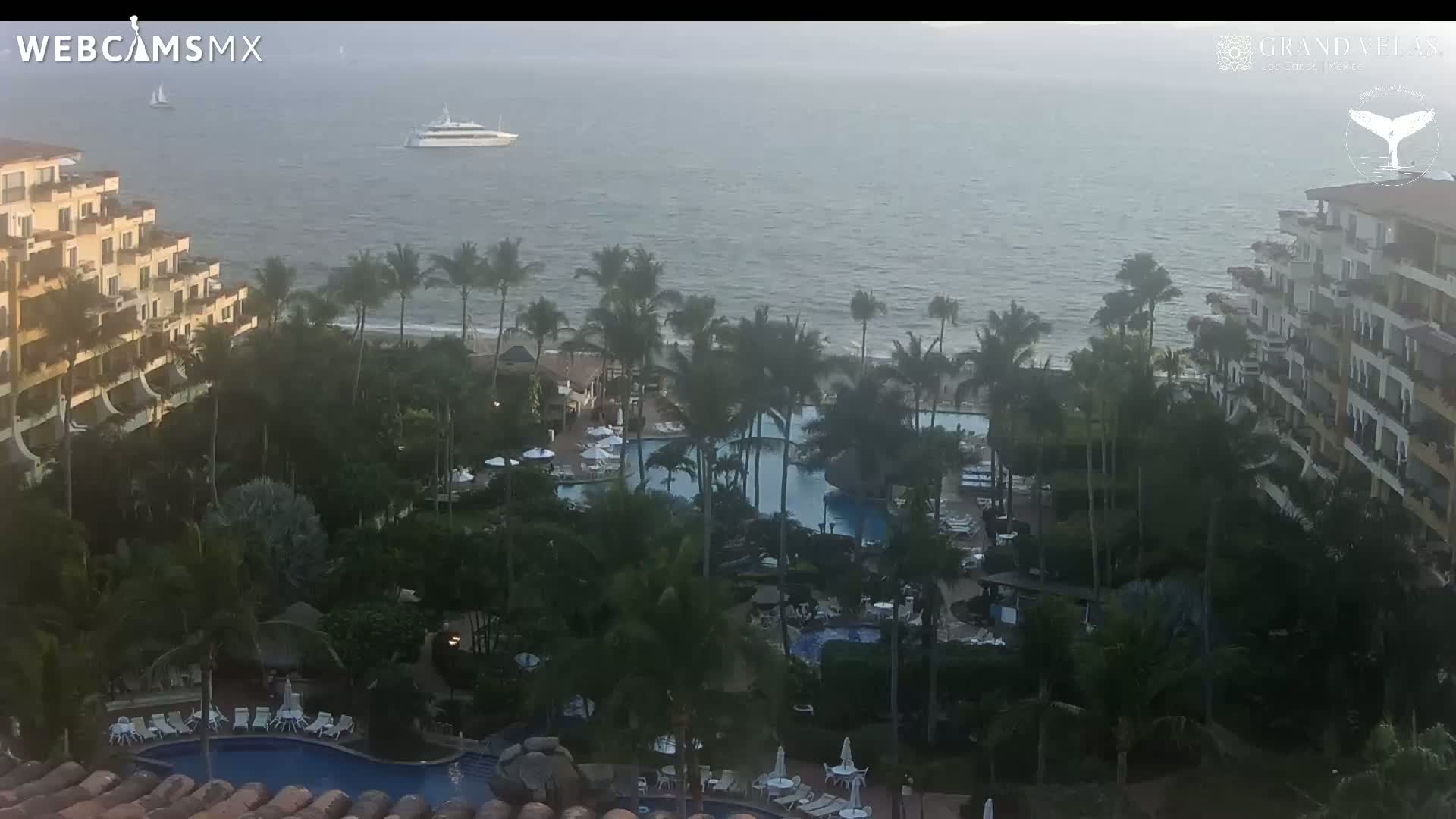 An aerial view shows a resort with multiple pools and palm trees situated on a coastline with calm ocean water and a large boat in the distance.