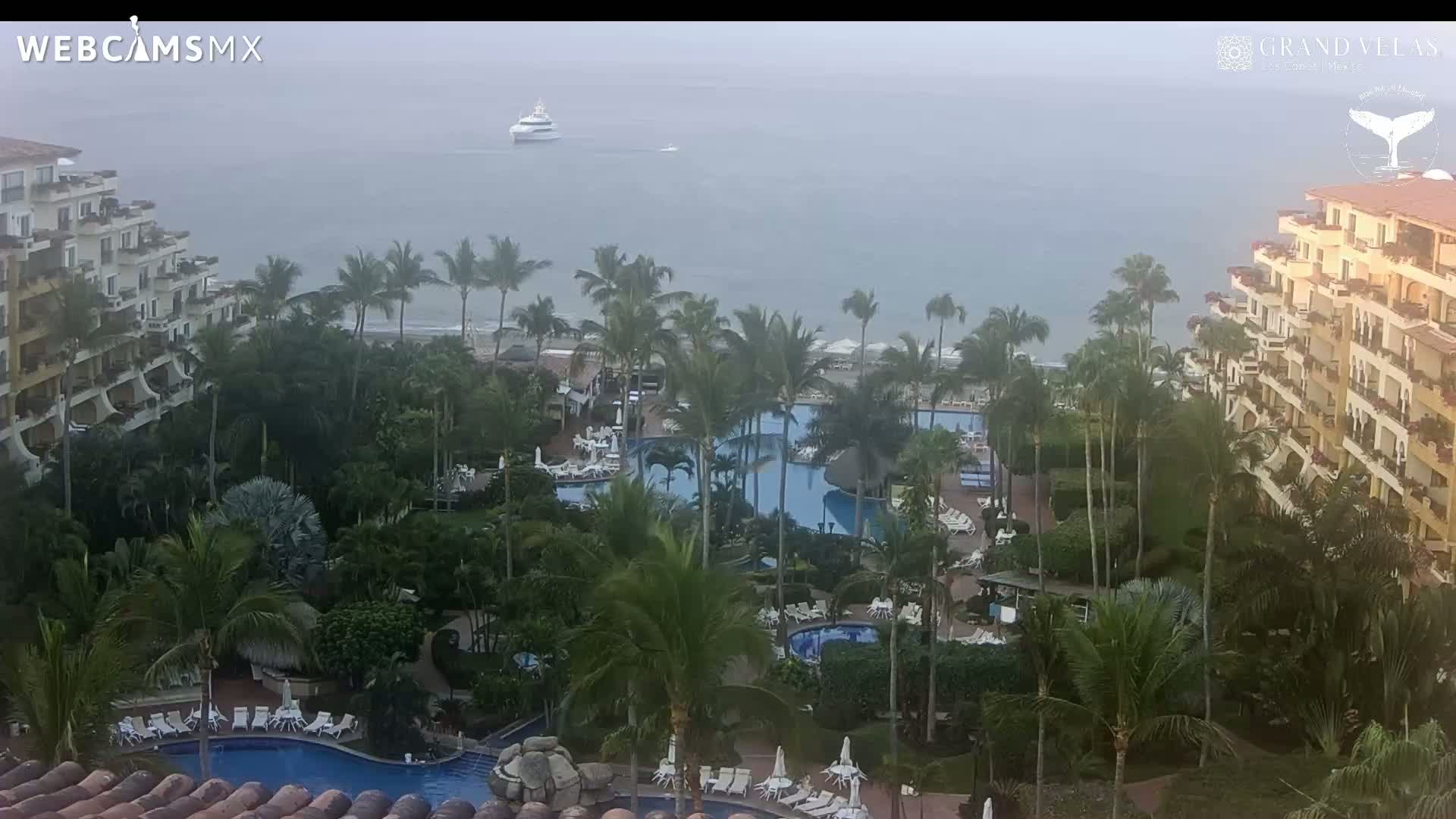 An aerial view of a tropical resort with multiple pools surrounded by palm trees and buildings, overlooking a hazy ocean with a distant boat.