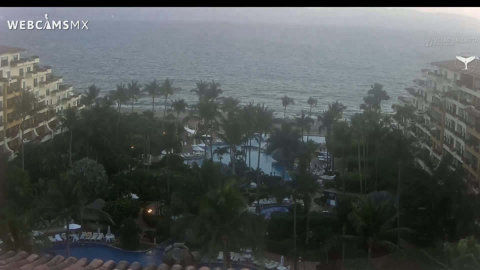 A resort with multiple pools surrounded by palm trees overlooks a somewhat choppy ocean under an overcast sky.