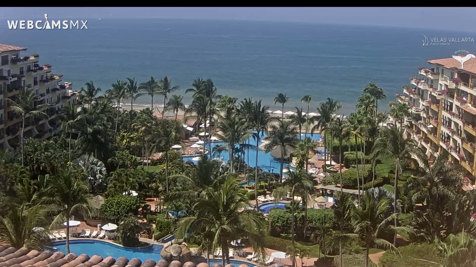 A tropical resort with multiple pools and palm trees overlooks a calm ocean on a sunny day.